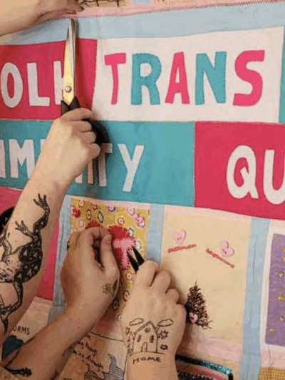 A person with tattooed arms can be seen hanging up a community quilt labelled "Norfolk Trans Joy Community Quilt."