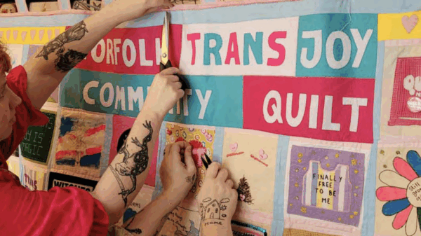 A person with tattooed arms can be seen hanging up a community quilt labelled "Norfolk Trans Joy Community Quilt."
