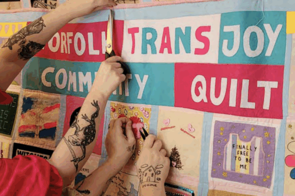A person with tattooed arms can be seen hanging up a community quilt labelled "Norfolk Trans Joy Community Quilt."