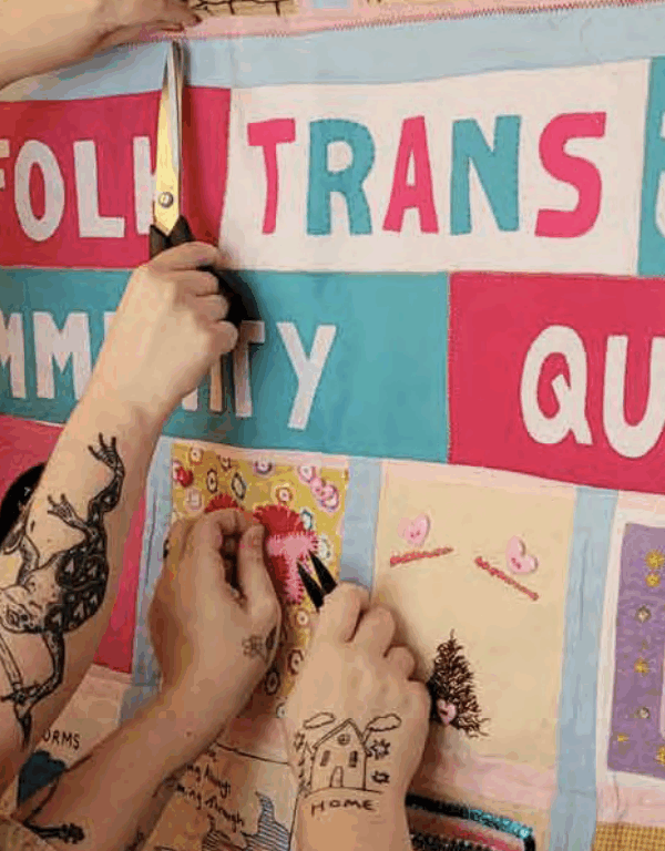 A person with tattooed arms can be seen hanging up a community quilt labelled "Norfolk Trans Joy Community Quilt."