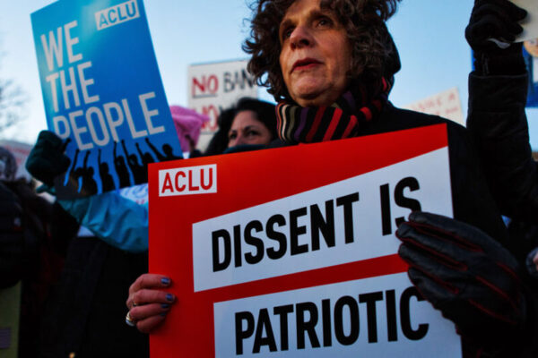 A white woman is in the crowd with a red sign that says "Dissent is Patriotic." Behind him is a blue sign that says, "WE THE PEOPLE."