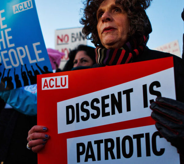 A white woman is in the crowd with a red sign that says "Dissent is Patriotic." Behind him is a blue sign that says, "WE THE PEOPLE."
