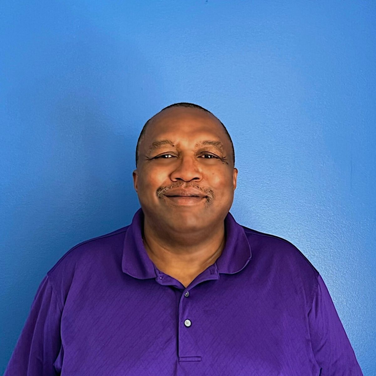 Anderson Curtis, a Black man wearing a purple shirt, smiles at the camera in front of a blue wall.