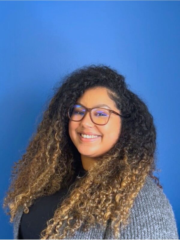 An Afro-Bori woman with curly hair, her roots are dark and her ends are bleached, in a grey cardigan smiles at the camera in front of a blue wall.