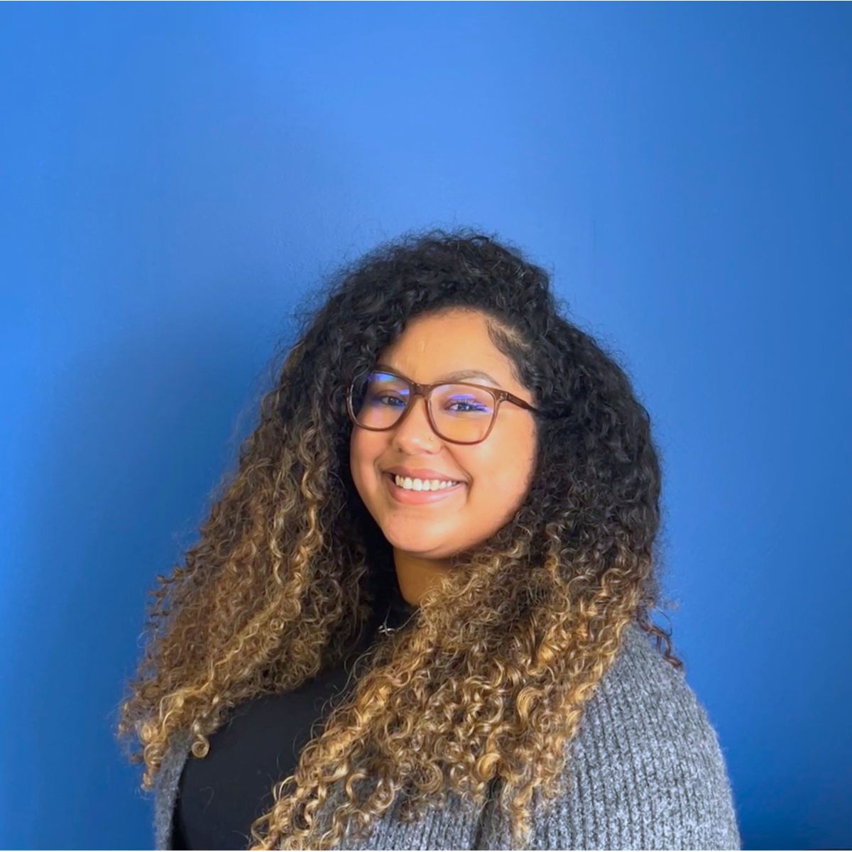 An Afro-Bori woman with curly hair, her roots are dark and her ends are bleached, in a grey cardigan smiles at the camera in front of a blue wall.