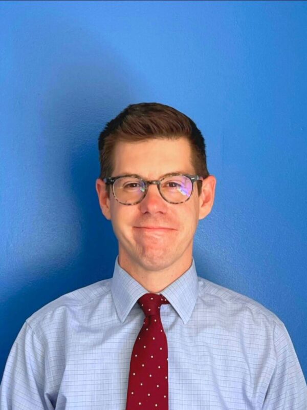 David McGuire, a white man with short brown hair wearing a light blue dress shirt with a red tie and brown glasses, looking at the camera with a smile.