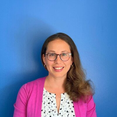 A woman with brown hair to her shoulders, swept over her left shoulder, with brown glasses and blue/green eyes smiles at the camera in front of a blue wall with a pink sweater on.