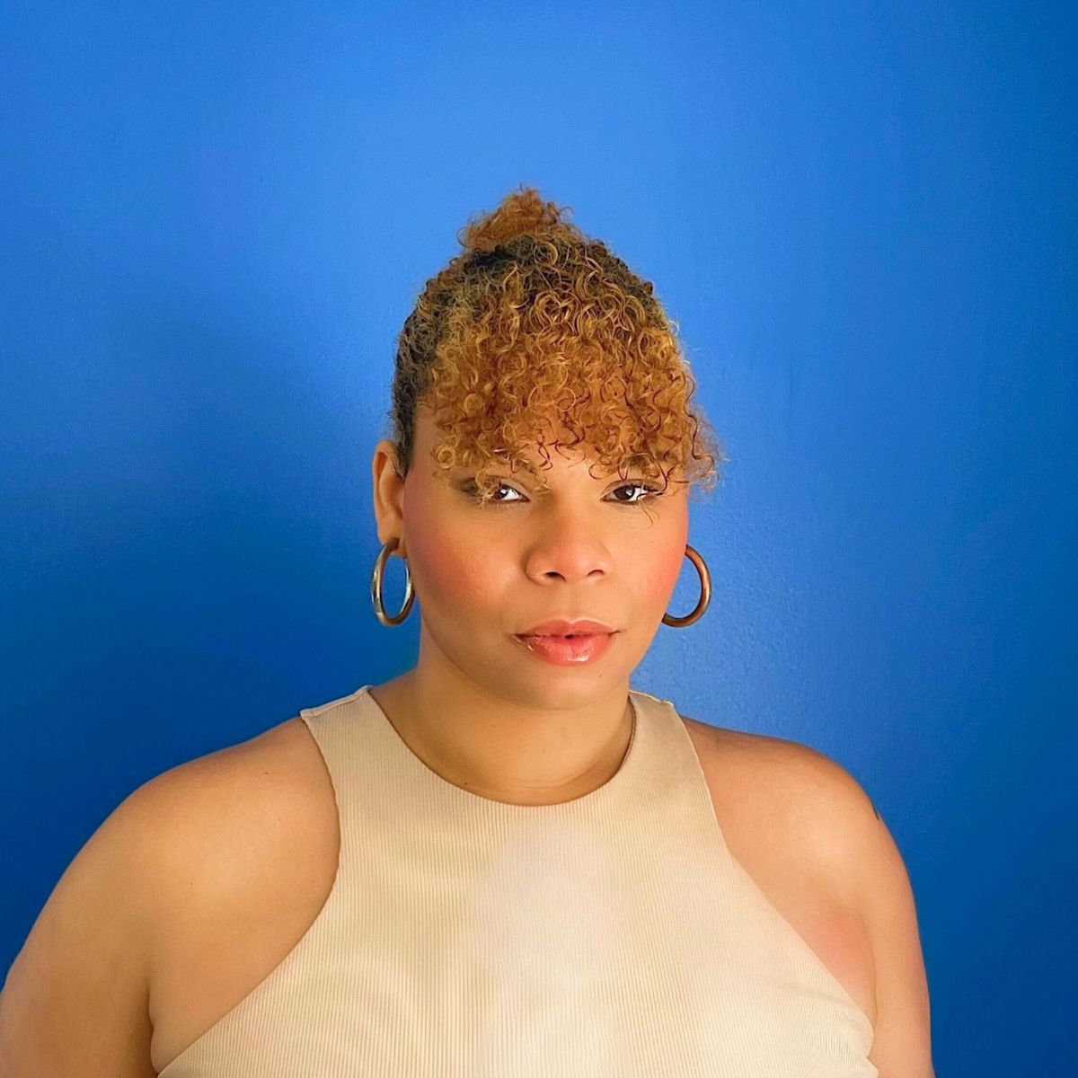A Black, Taíno woman looks at the camera with her curled hair up in a claw clip, wearing gold hoops and a nude-colored top in front of a blue wall.