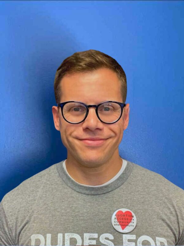 Gus Marks-Hamilton, a white man with black round glasses, smiles at the camera in front a blue wall with a gray shirt that says "Dudes for Abortions."