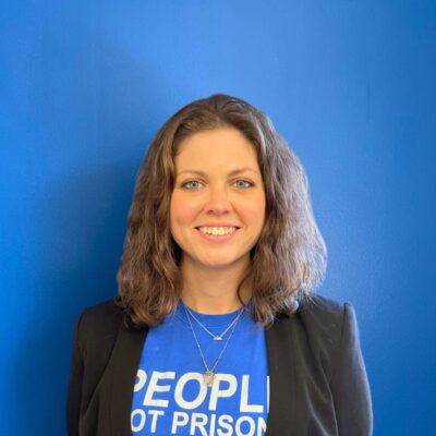 A white woman with light brown hair to her shoulders smiles at the camera in a blue People not Prisons t-shirt and blazer combo, standing in front of a blue wall.