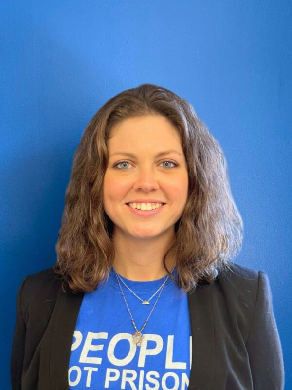 A white woman with light brown hair to her shoulders smiles at the camera in a blue People not Prisons t-shirt and blazer combo, standing in front of a blue wall.