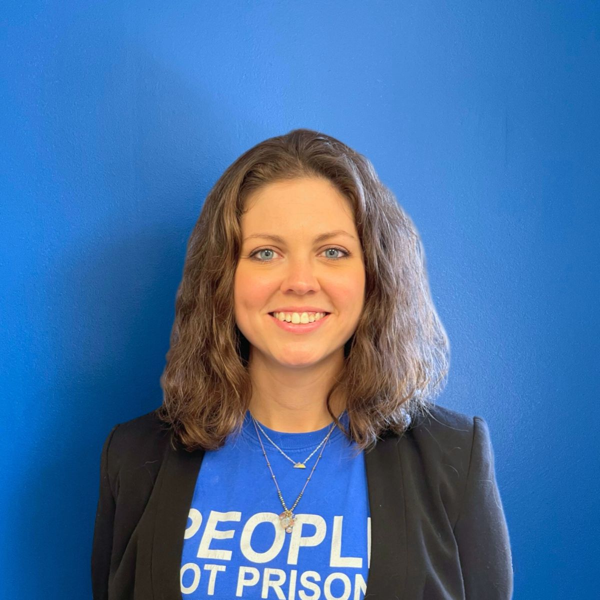 A white woman with light brown hair to her shoulders smiles at the camera in a blue People not Prisons t-shirt and blazer combo, standing in front of a blue wall.