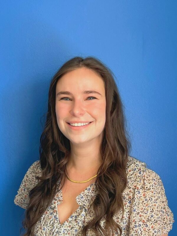 Jess Zaccagnino, a white woman wearing a floral dress with long brown hair past her shoulders, smiling at the camera in front of a blue wall.