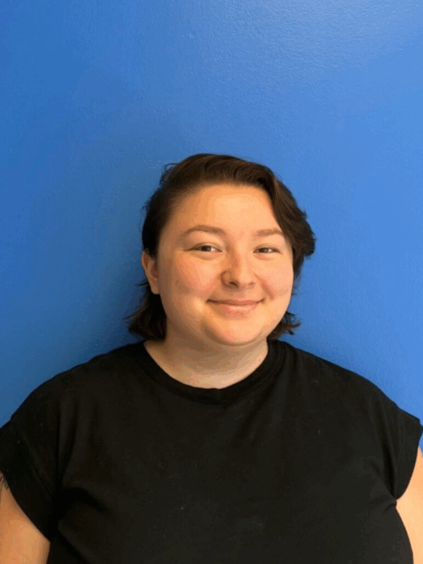 On a blue background, a person looks at the camera with a smile. Kira is wearing a black t-shirt and has short, dark hair with bangs on their left side.