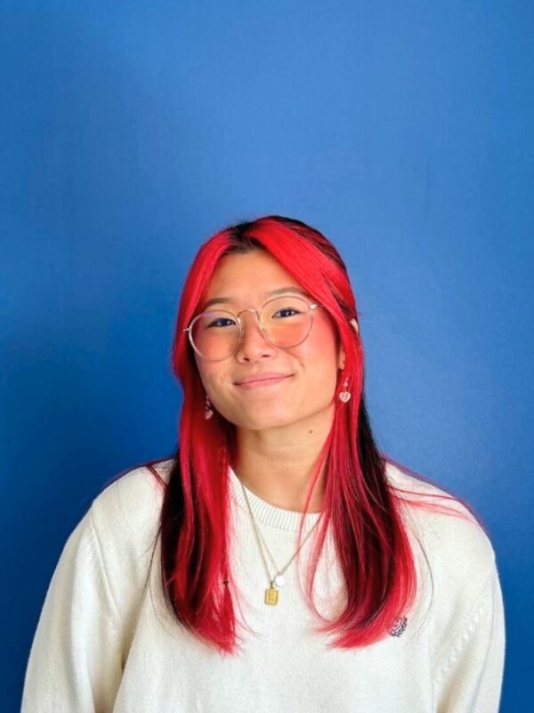 A Korean woman with long black hair, with red dyed hair in the front of and under her black hair is smiling at the camera. She is wearing gold-rim glasses and a white sweater on a blue background.