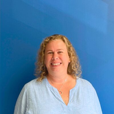 A white woman with curly blonde hair to her shoulders in a collared shirt smiles at the camera in front of a blue wall.