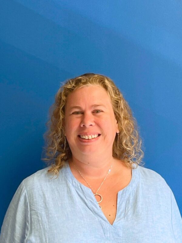 A white woman with curly blonde hair to her shoulders in a collared shirt smiles at the camera in front of a blue wall.