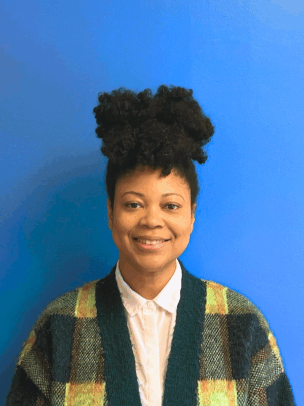 A Black woman with her curls up on top of her head in a green plaid cardigan smiles at the camera in front of a blue wall.