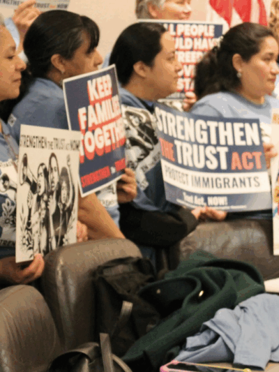 A group of women are standing in a hearing room at the Capitol, holding signs that says "Undocumented + Unafraid" and "Strengthen the Trust Act Now!"