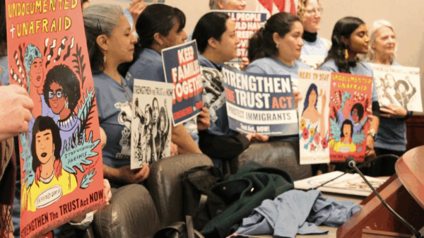 A group of women are standing in a hearing room at the Capitol, holding signs that says "Undocumented + Unafraid" and "Strengthen the Trust Act Now!"