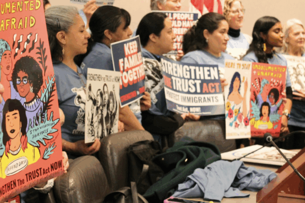 A group of women are standing in a hearing room at the Capitol, holding signs that says "Undocumented + Unafraid" and "Strengthen the Trust Act Now!"