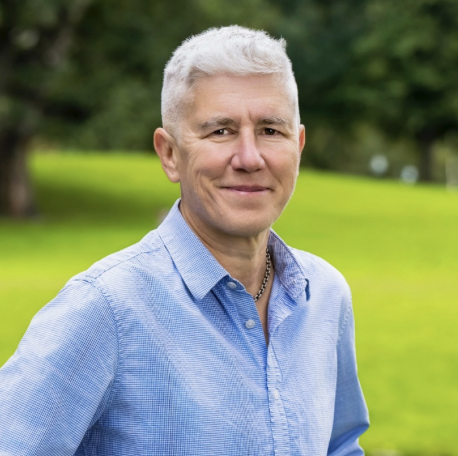 Jennifer Levi, a white woman with short white hair, is smiling at the camera wearing a blue button up and standing in front of a grassy hill.