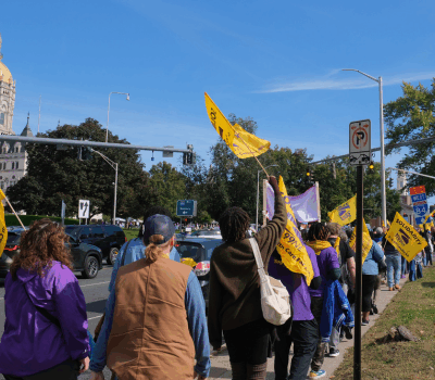 Walking towards the Capitol in Hartford is a group of members of the SEIU union. Several folks are identifiable in their purple union t-shirts, holding big yellow flags with the union logo on them.