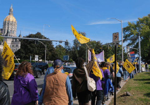 Walking towards the Capitol in Hartford is a group of members of the SEIU union. Several folks are identifiable in their purple union t-shirts, holding big yellow flags with the union logo on them.