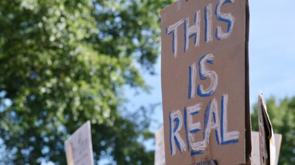In the focal point of the picture, on the right of the picture, is a brown cardboard sign being held up by a stick that reads in white: "THIS IS REAL"