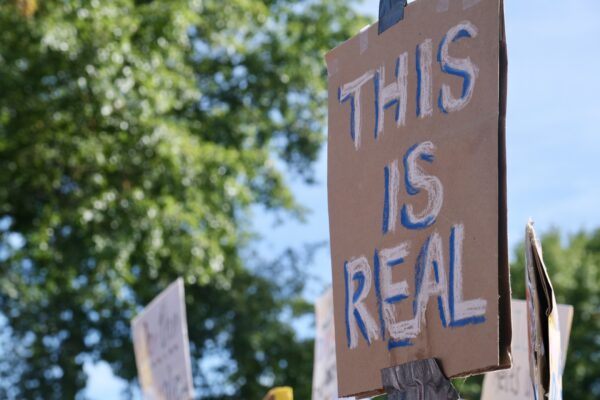 In the focal point of the picture, on the right of the picture, is a brown cardboard sign being held up by a stick that reads in white: "THIS IS REAL"