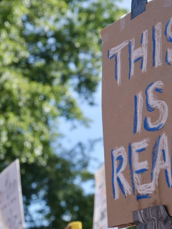 In the focal point of the picture, on the right of the picture, is a brown cardboard sign being held up by a stick that reads in white: "THIS IS REAL"
