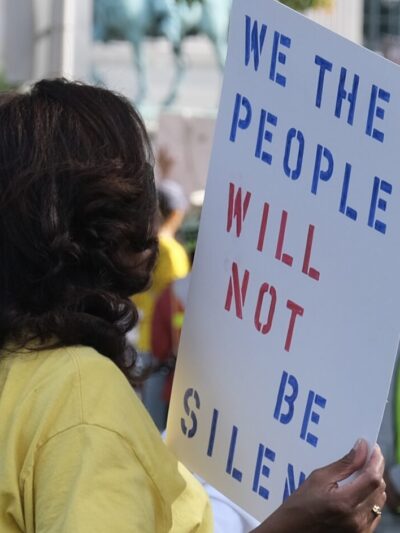 A woman in a crowd is holding a sign that says: "We the people will not be silenced."
