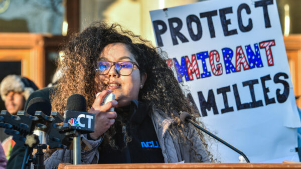 Standing behind a podium and in front of a white sign that reads "PROTECT IMMIGRANTS FAMILIES," our policy director Chelsea-Infinity is holding a microphone and speaking to the crowd in front of her (not pictured).