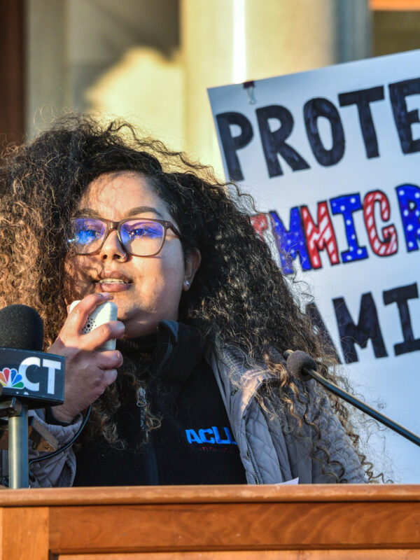 Standing behind a podium and in front of a white sign that reads "PROTECT IMMIGRANTS FAMILIES," our policy director Chelsea-Infinity is holding a microphone and speaking to the crowd in front of her (not pictured).