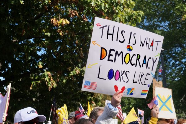 In front of a green tree, a white hand is holding up a white sign that reads: "THIS IS WHAT DEMOCRACY LOOKS LIKE"