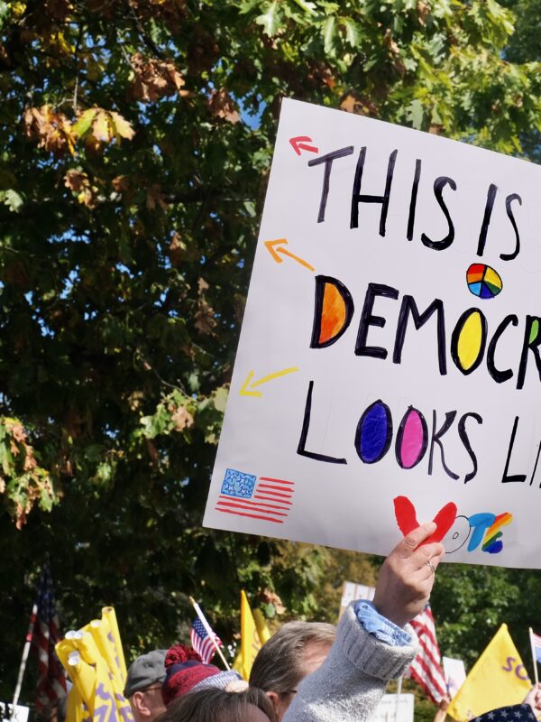 In front of a green tree, a white hand is holding up a white sign that reads: "THIS IS WHAT DEMOCRACY LOOKS LIKE"