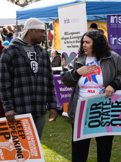 Cris, our community engagement program director, is speaking with a Black man in a flannel in the grass. Both are holding ACLU-branded signs that are orange and blue.
