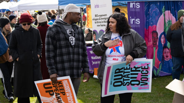 Cris, our community engagement program director, is speaking with a Black man in a flannel in the grass. Both are holding ACLU-branded signs that are orange and blue.