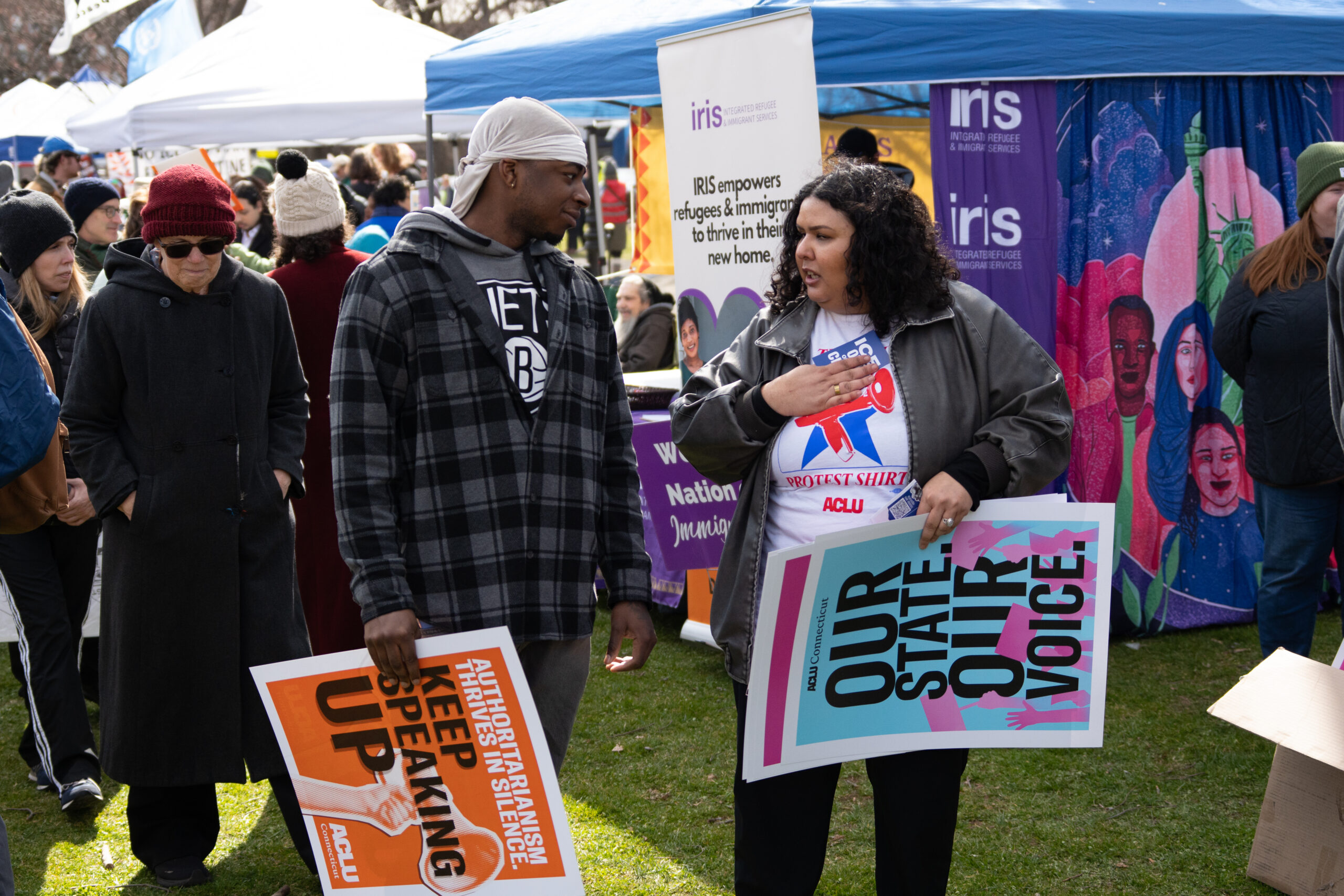Cris, our community engagement program director, is speaking with a Black man in a flannel in the grass. Both are holding ACLU-branded signs that are orange and blue.