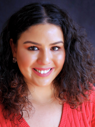 Cristher, our community engagement and education program director, smiles at the camera in front of a dark background wearing a pink v-neck sweater.
