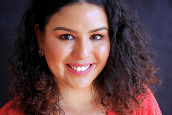 Cristher, our community engagement and education program director, smiles at the camera in front of a dark background wearing a pink v-neck sweater.