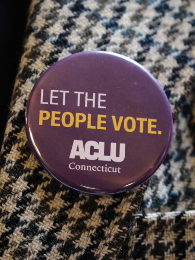 On a houndstooth jacket, our purple pin with white text that reads "Let the People Vote." with the white ACLU Connecticut logo beneath the text is in the center of the photo.