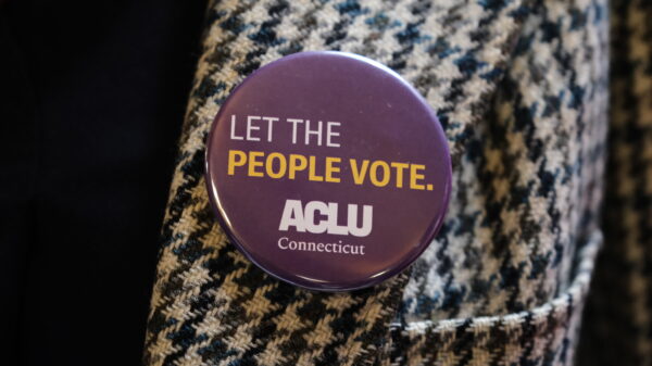 On a houndstooth jacket, our purple pin with white text that reads "Let the People Vote." with the white ACLU Connecticut logo beneath the text is in the center of the photo.