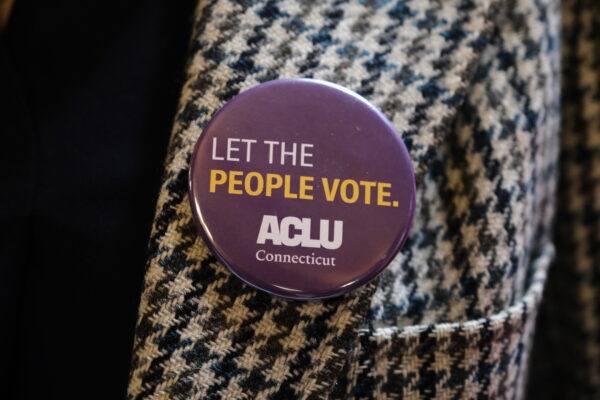 On a houndstooth jacket, our purple pin with white text that reads "Let the People Vote." with the white ACLU Connecticut logo beneath the text is in the center of the photo.