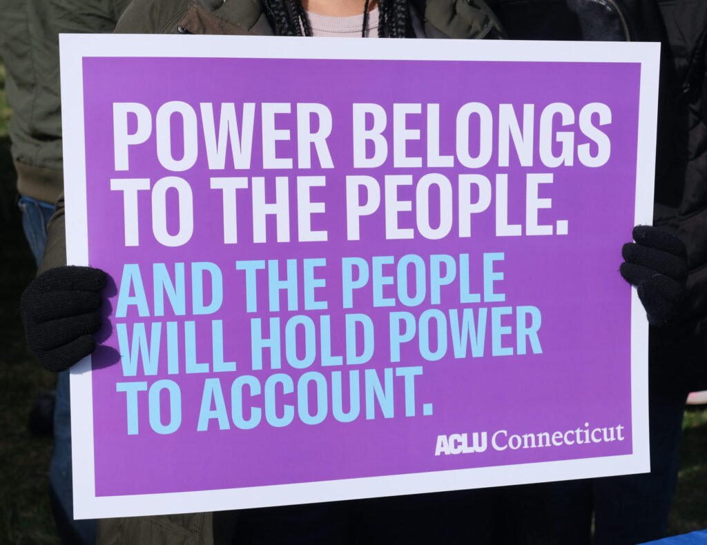 Zoomed in picture of a purple sign that says "Power belongs to the people." in white and below that text is "And the people will hold power to account." in blue.