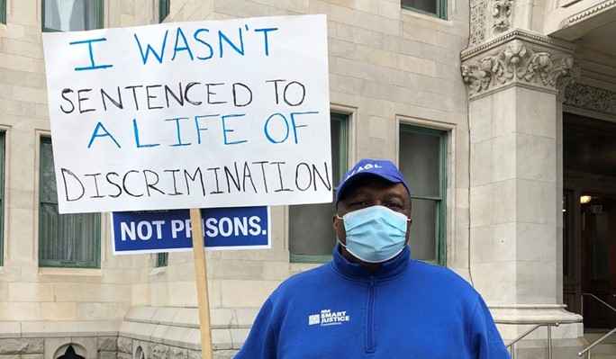 Anderson Curtis, in blue sweatshirt and hat and mask, holds a sign that says "I wasn't sentenced to a life of discrimination"