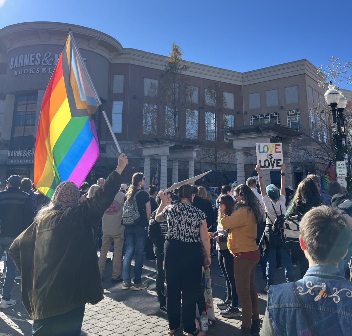 In front of a Barnes & Nobles, a crowd stands in front of a stage. Several people are holding signs, LGBTQIA+ flags, etc.