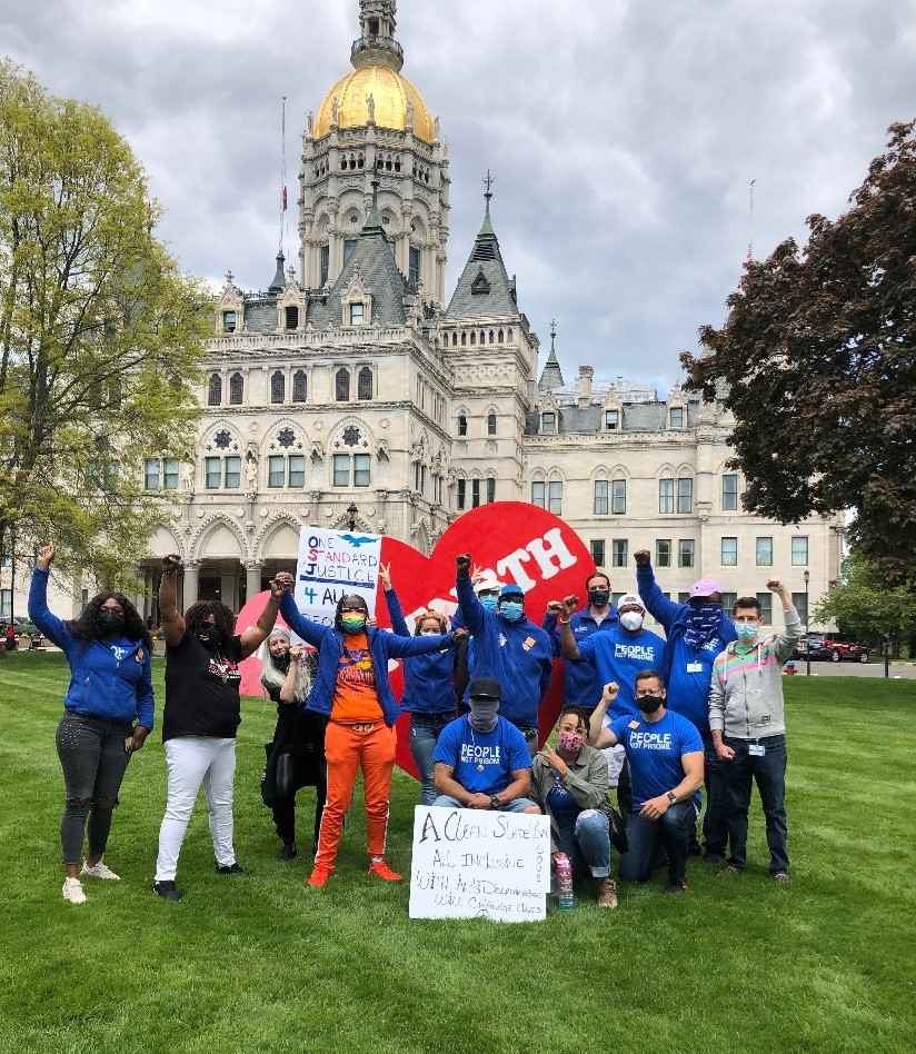 ACLUCT Smart Justice leaders, in blue people not prisons shirts, crouch and stand with a massive red heart that says "strength" in white letters. Behind them is the CT capitol building. They are masked and raising their fists in celebration and power.