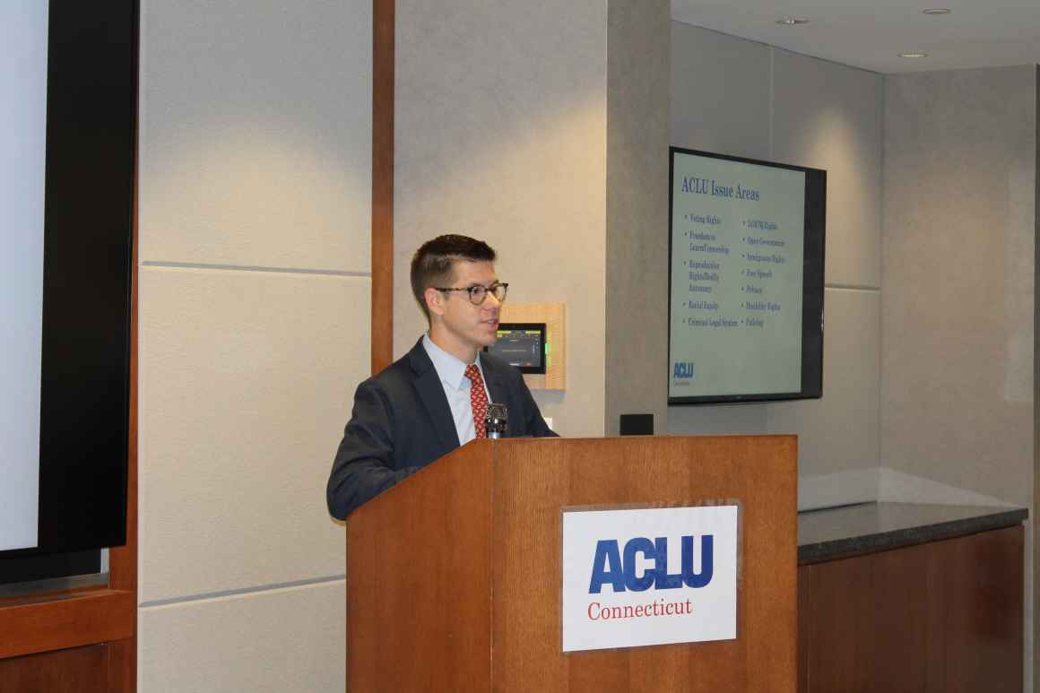 A white man with dark hair and glasses stands behind a wooden podium that has the ACLU-CT logo on it.