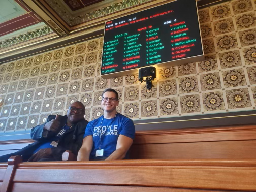 Anderson Curtis and Gus Marks-Hamilton sit, smiling, with Anderson giving a thumbs up, in front of the vote board for CT's 2023 prosecutorial accountability law.